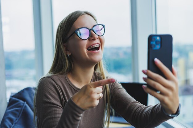 Young smiling woman wearing glasses in casual clothes sitting\
in a chair by the window and talking on the phone during a video\
call