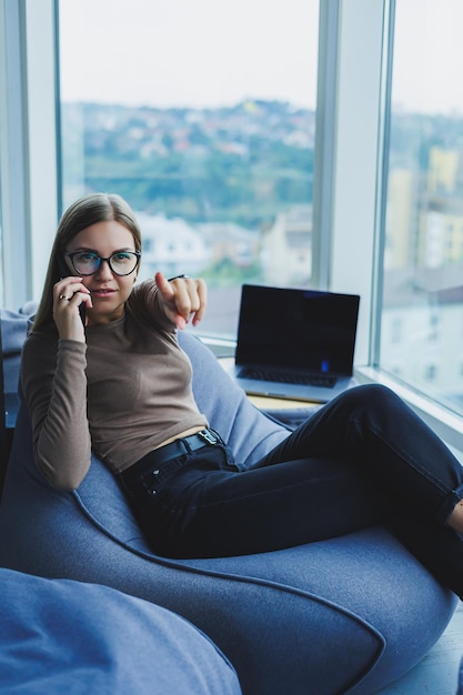 Young smiling woman wearing glasses in casual clothes sitting\
in a chair by the window and talking on the phone during a video\
call