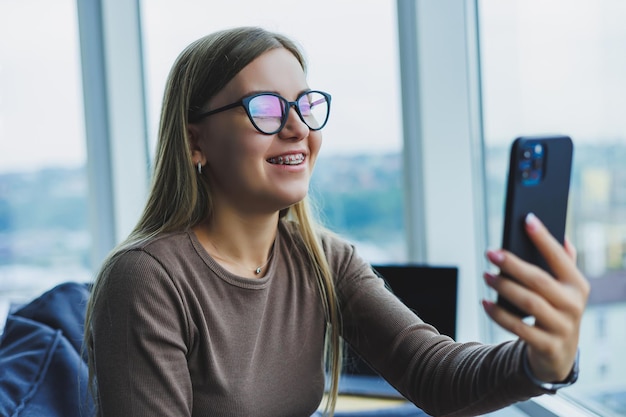 窓際の椅子に座って、ビデオ通話中に電話で話しているカジュアルな服装で眼鏡をかけている若い笑顔の女性