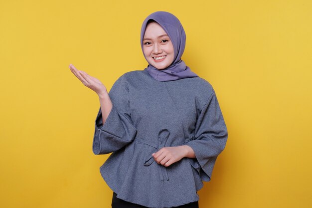 Young smiling woman wearing hijab shows something and holding something with palms isolated on light yellow banner background