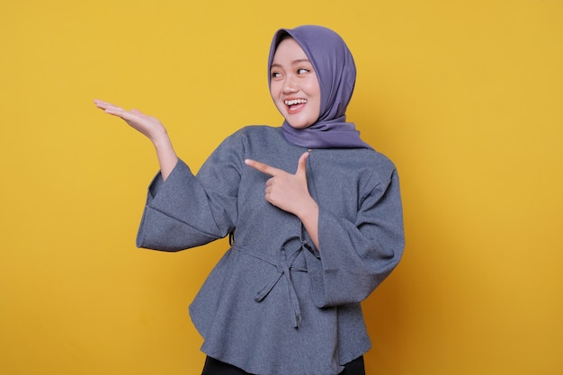 Young smiling woman wearing hijab shows something and holding something with palms isolated on light yellow banner background