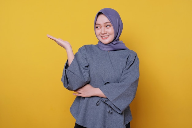 Young smiling woman wearing hijab shows something and holding something with palms isolated on light yellow banner background