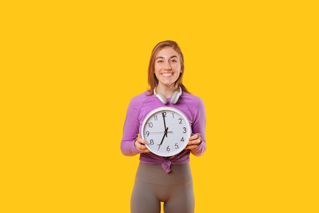 Young smiling woman wearing a pair of headphones is holding a clock