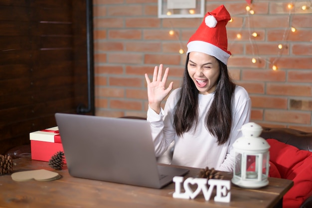 A young smiling woman wearing red Santa Claus hat making video call on social network with family and friends on Christmas day.