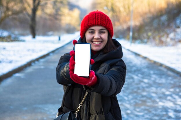 Young smiling woman in winter outfit holding phone with white blank empty screen