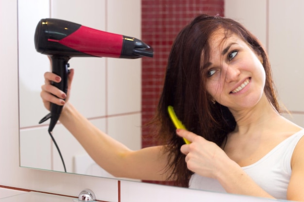 Young smiling woman with hairdryer and yellow comb in front of the mirror in her bathroom