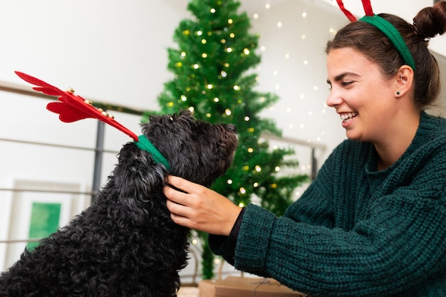 クリスマス休暇中にトナカイの角を身に着けている彼女の黒いふわふわ犬と若い笑顔の女性