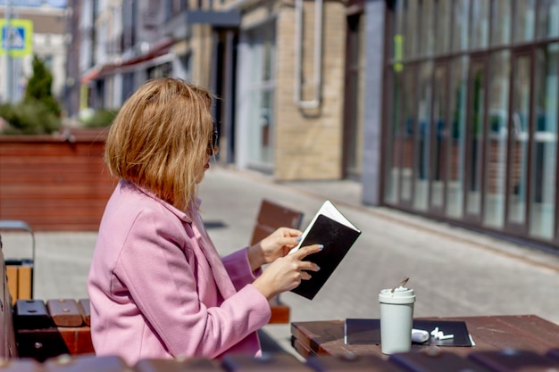 A young smiling womangirl reads a book resting on a bench with coffee against the background of a bustling city