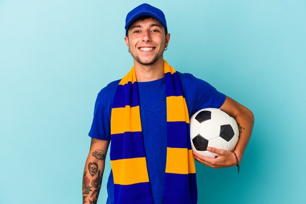 Young soccer fan man holding a ball isolated on blue background  happy, smiling and cheerful.