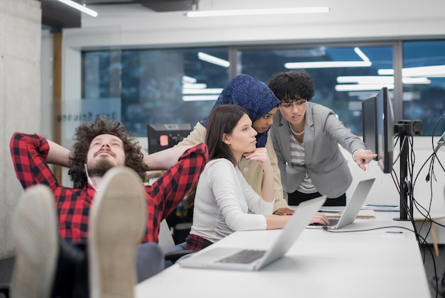 young software developer resting with legs on desk while his multiethnics business team writing programming code in the background at startup office