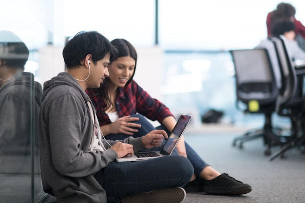 若いソフトウェア開発者は、現代の創造的なスタートアップオフィスの床に座ってプログラミングコードを書くラップトップコンピューターを使用してカップル