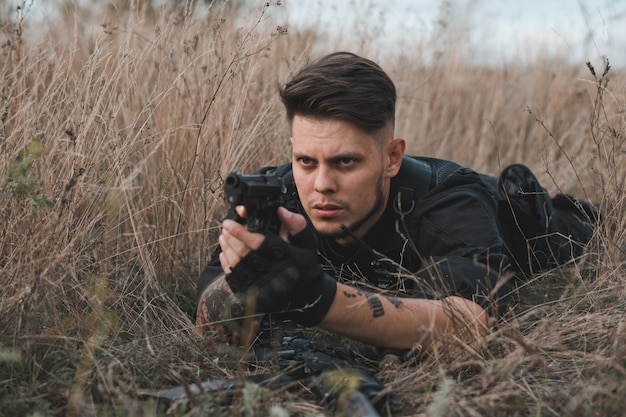 Young soldier in black uniform lying down and aiming a pistol