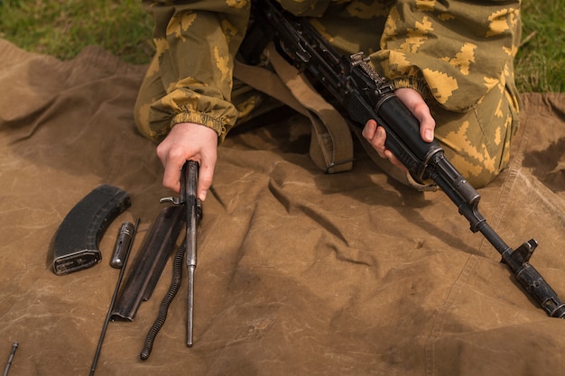 迷彩服を着た若い兵士が武器を解体しています。男は軍事演習で芝生の上でカラシニコフ突撃ライフルを収集します。