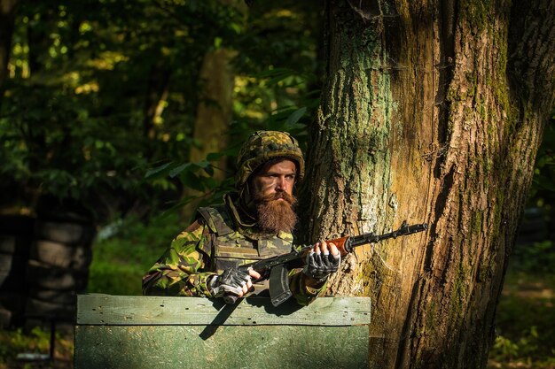 Young soldier hipster with beard on dirty tired face in military ammunition and helmet standing on guard near tree and wooden board with gun in hands in forest