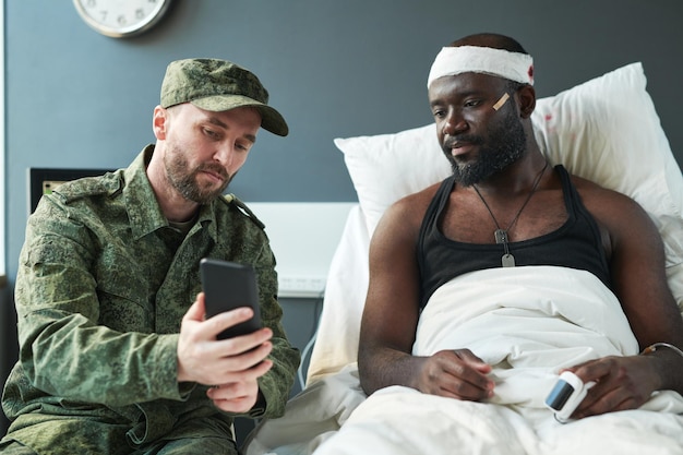Young soldier in military uniform showing new photos in smartphone to his friend