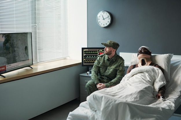 Young soldier in military uniform sitting by bed of injured friend