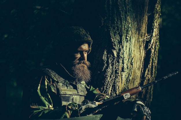 Молодой солдат с бородой на грязном усталом лице в военной форме и шлеме с винтовкой на дереве и г