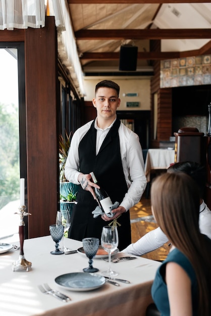 A young sommelier in a stylish apron demonstrates and offers fine wine to a beautiful couple in a restaurant Customer service in an elite restaurant and a public restaurant