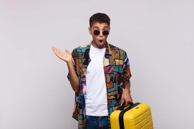 Young south american man looking surprised and shocked, with jaw dropped holding an object with an open hand on the side