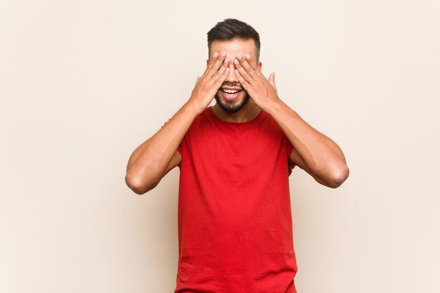 Young south-asian man covers eyes with hands, smiles broadly waiting for a surprise.