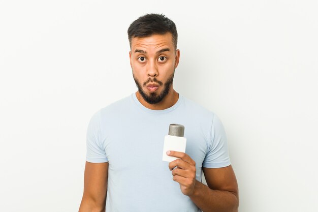 Young south-asian man holding an after shave cream shrugs shoulders and open eyes confused.