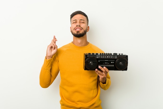 Young south-asian man holding a guetto blaster crossing fingers for having luck