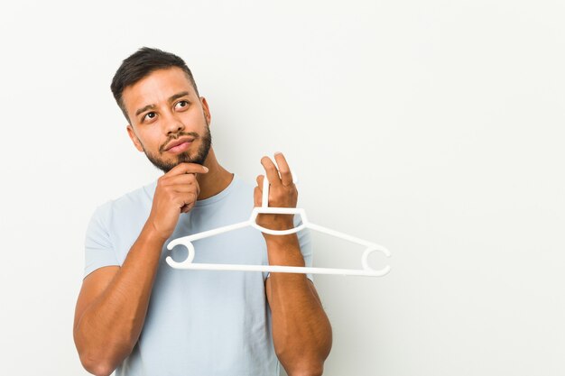 Young south asian man holding a hanger looking sideways with doubtful and skeptical expression.