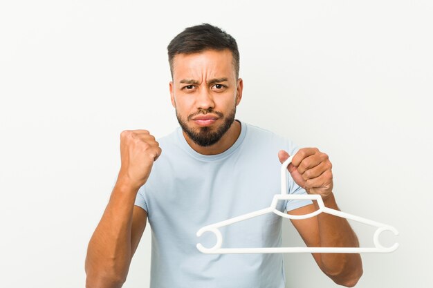 Young south asian man holding a hanger showing fist to with aggressive facial expression.