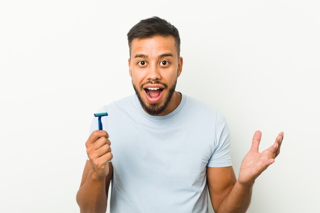 Young south-asian man holding a razor blade receiving a pleasant surprise, excited and raising hands.