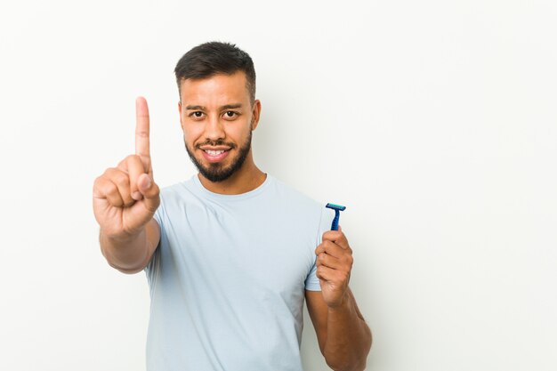 Young south-asian man holding a razor blade showing number one with finger.