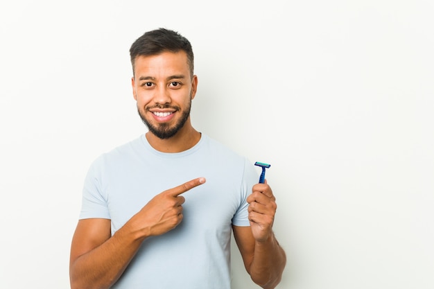 Young south-asian man holding a razor blade smiling and pointing aside, showing something