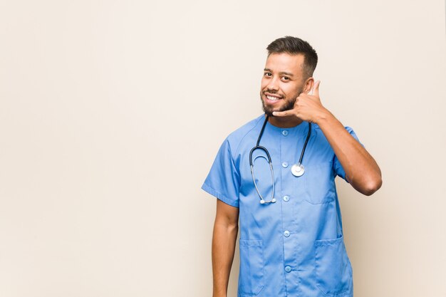Young south-asian nurse man showing a mobile phone call gesture with fingers.