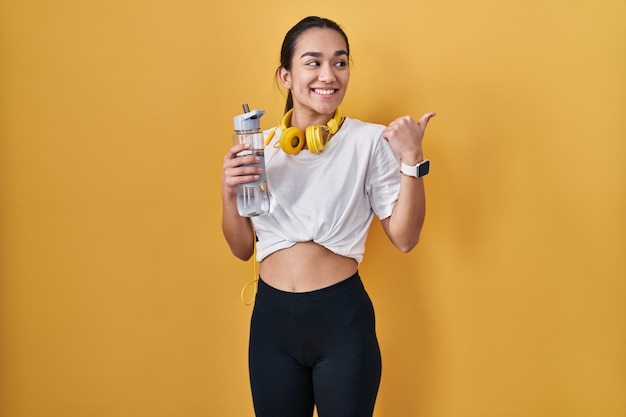 Young south asian woman wearing sportswear drinking water smiling with happy face looking and pointing to the side with thumb up