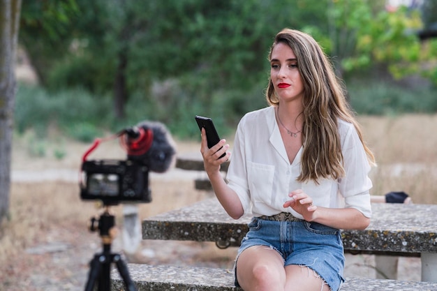 young spanish female blogger using her phone and recording herself with a dslr camera in a park