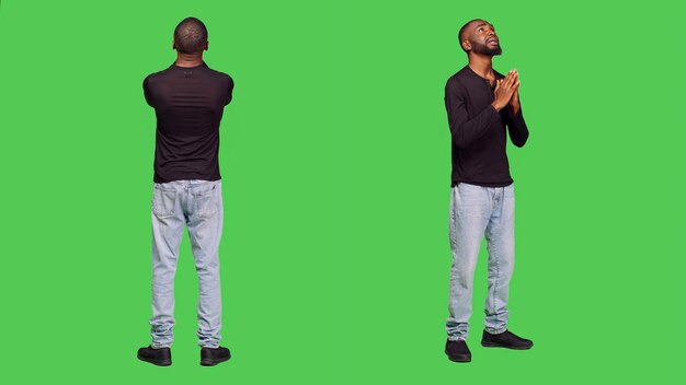 Young spiritual man praying to jesus in studio, holding hands in a prayer and asking for forgiveness. Person being religious with belief, optimistic adult on full body green screen backdrop.