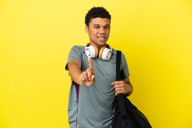 Young sport African American man with sport bag isolated on yellow background showing and lifting a finger