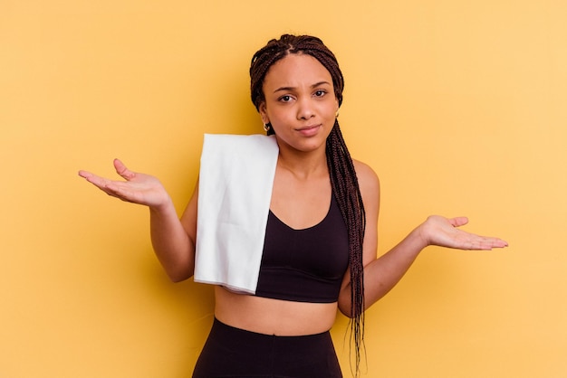 Young sport donna afro-americana che tiene un asciugamano isolato su sfondo giallo dubitando e scrollando le spalle in gesto interrogativo.