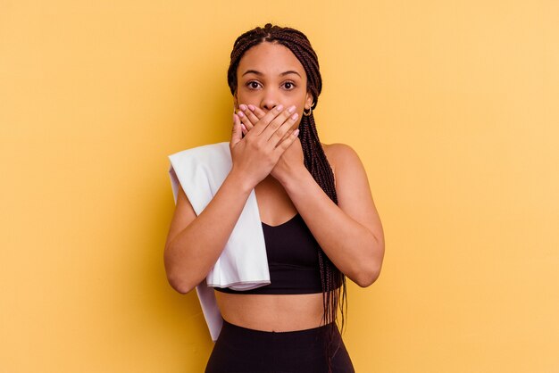 Young sport african american woman holding a towel isolated on yellow background shocked covering mouth with hands.