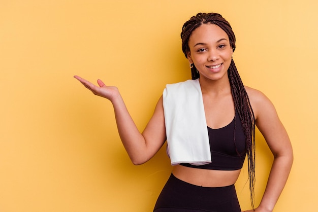 Young sport african american woman holding a towel isolated on yellow background showing a copy space on a palm and holding another hand on waist.