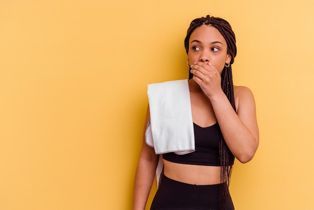 Young sport african american woman holding a towel isolated on yellow wall thoughtful looking to a copy space covering mouth with hand