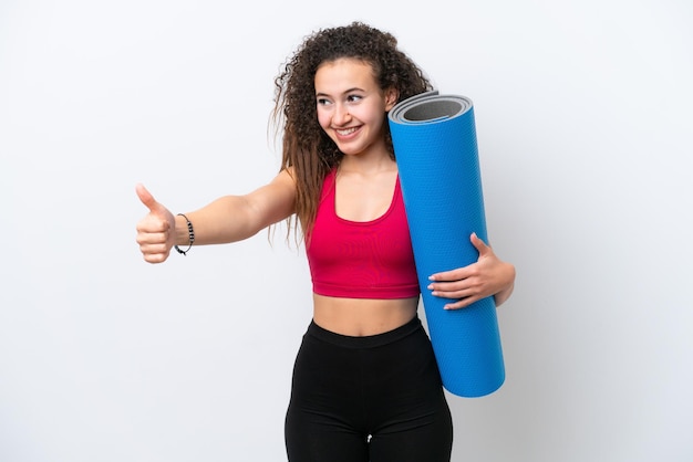 Young sport Arab woman going to yoga classes while holding a mat isolated on white background giving a thumbs up gesture