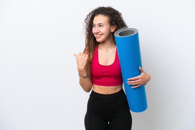 Young sport Arab woman going to yoga classes while holding a mat isolated on white background pointing to the side to present a product