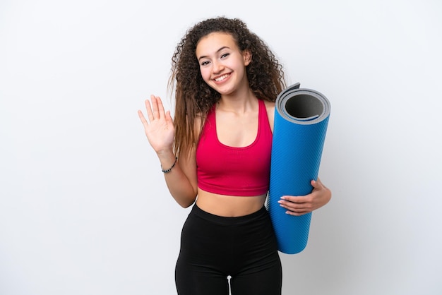 Young sport Arab woman going to yoga classes while holding a mat isolated on white background saluting with hand with happy expression