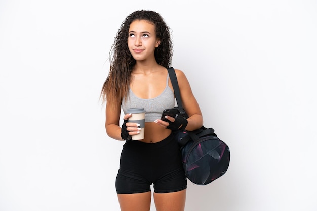 Young sport arab woman with sport bag isolated on white\
background holding coffee to take away and a mobile while thinking\
something