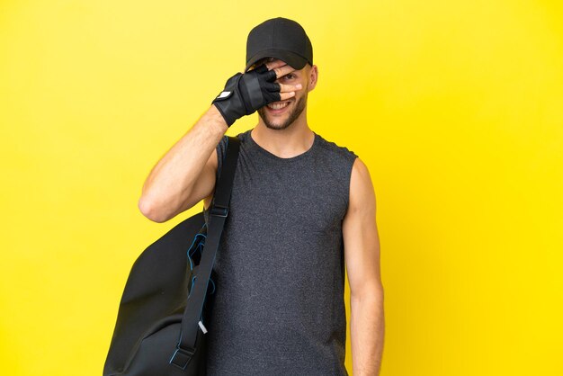 Young sport blonde man with sport bag isolated on yellow background covering eyes by hands and smiling