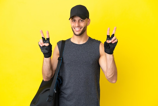 Young sport blonde man with sport bag isolated on yellow background showing victory sign with both hands