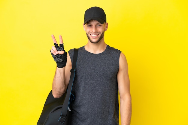 Young sport blonde man with sport bag isolated on yellow background smiling and showing victory sign