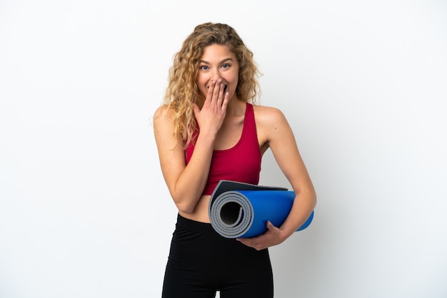 Young sport blonde woman going to yoga classes while holding a mat isolated on white background happy and smiling covering mouth with hand