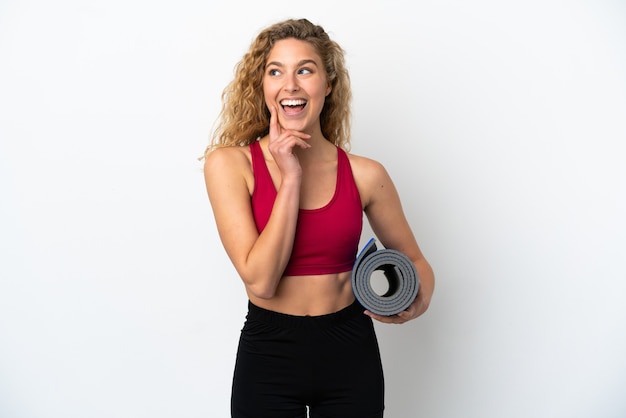 Giovane donna bionda sportiva che va a lezioni di yoga mentre tiene in mano un tappetino isolato su sfondo bianco pensando a un'idea mentre guarda in alto