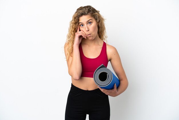 Giovane donna bionda sportiva che va alle lezioni di yoga mentre tiene in mano un tappetino isolato su sfondo bianco pensando a un'idea